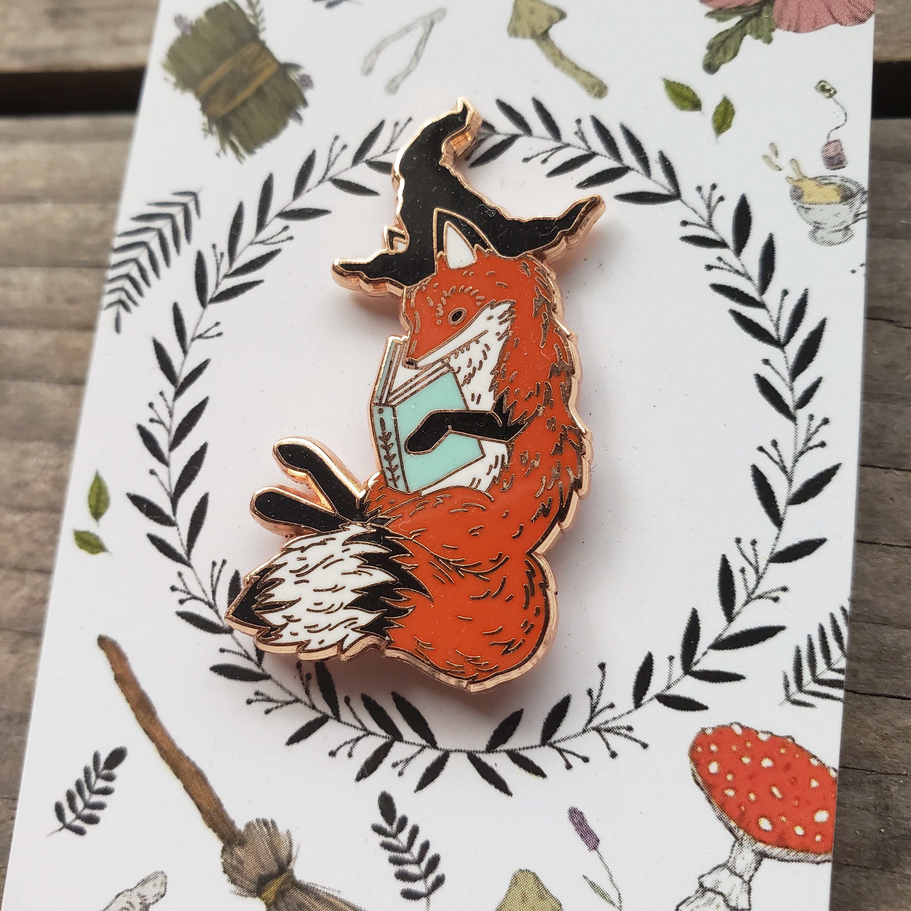 Enamel pin of a magical fox in a witch hat, reading a book.